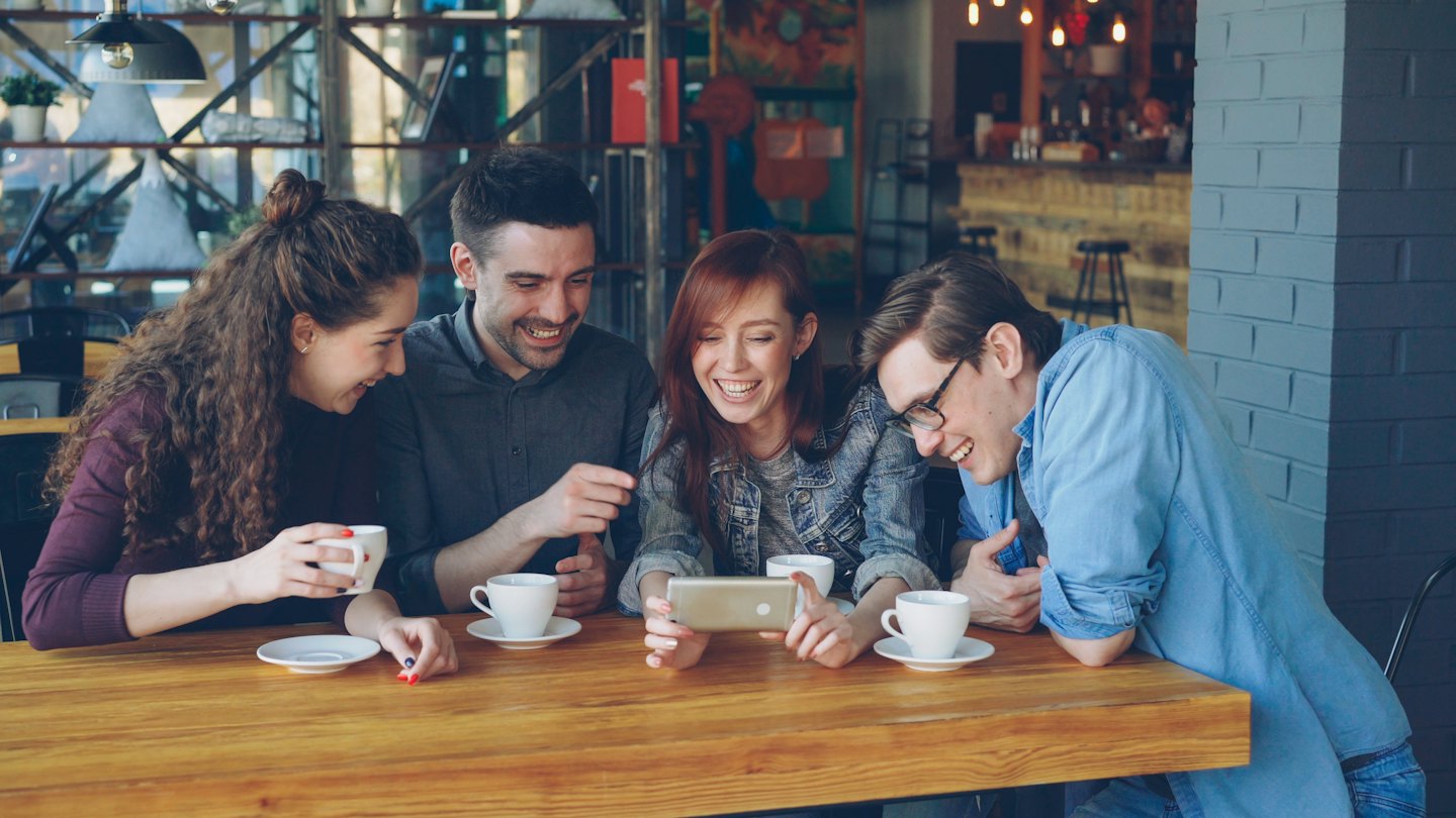Four friends laugh at a smartphone at a cafe table.